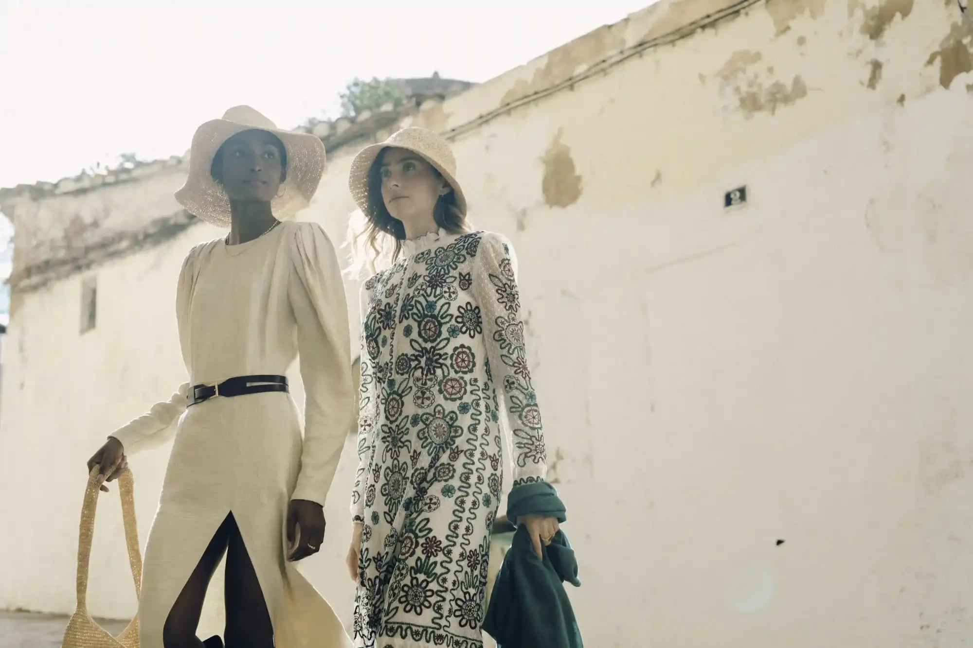 The woman on the right wears a white dress with intricate black floral embroidery and long sleeves, paired with a matching wide-brimmed straw hat.